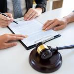 Close-up of three people’s hands at a desk, two wedding rings on a document, one person signing, another pointing at the paper, with a gavel and sound block in the foreground, suggesting a legal or divorce proceeding.