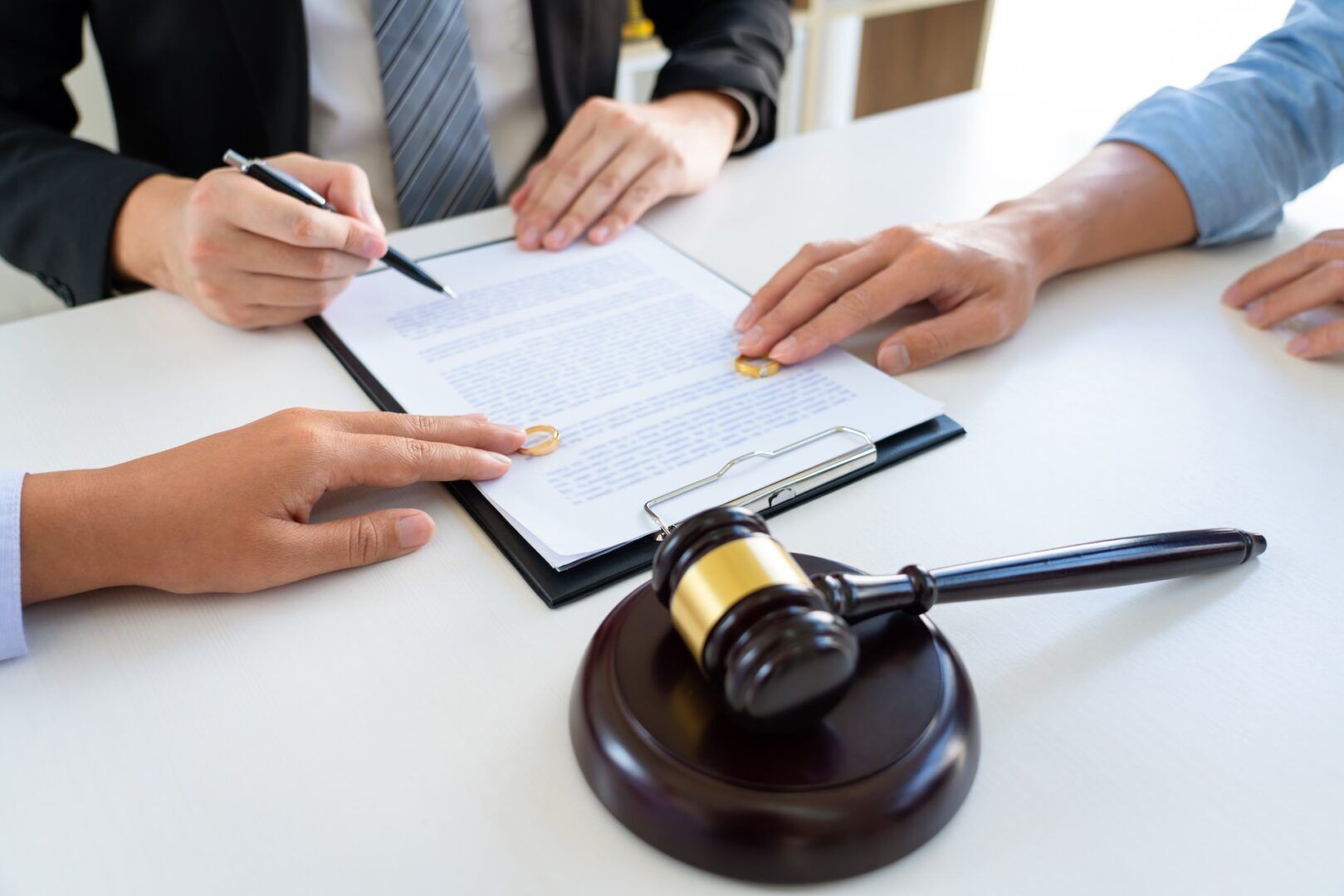 Close-up of three people’s hands at a desk, two wedding rings on a document, one person signing, another pointing at the paper, with a gavel and sound block in the foreground, suggesting a legal or divorce proceeding.
