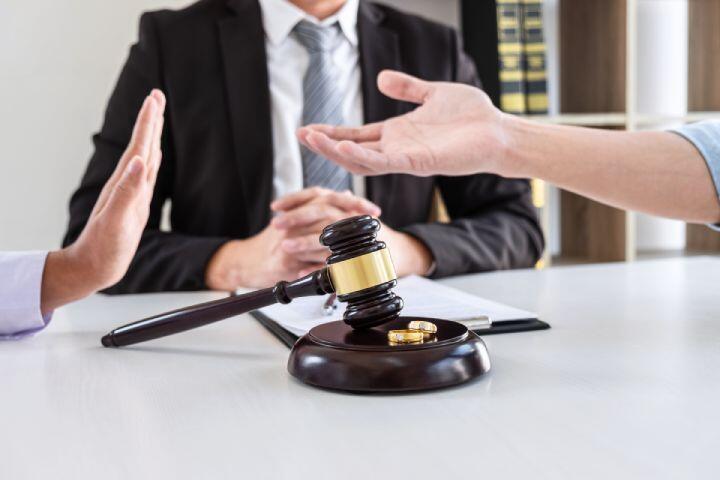 A judge sits at a desk with a gavel and two wedding rings, as two people gesture with their hands, suggesting a discussion or disagreement, possibly about divorce or legal separation.
