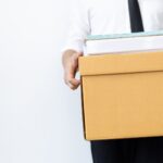 A person in business attire holds a cardboard box filled with office supplies and documents, standing against a plain white background.