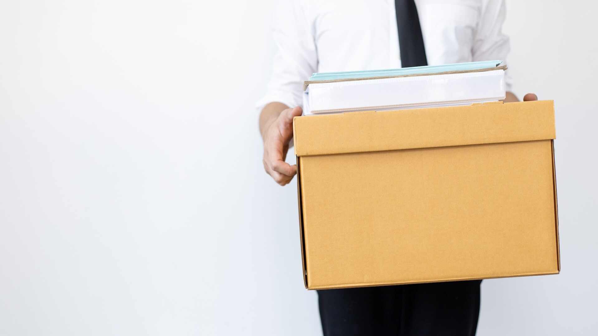 A person in business attire holds a cardboard box filled with office supplies and documents, standing against a plain white background.