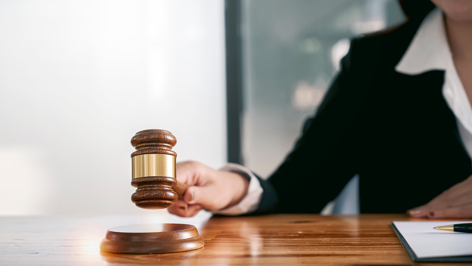 A person in a suit is holding a wooden gavel above a sound block on a desk, suggesting a legal or courtroom setting. A person in a suit is holding a wooden gavel above a sound block on a desk, suggesting a legal or courtroom setting.