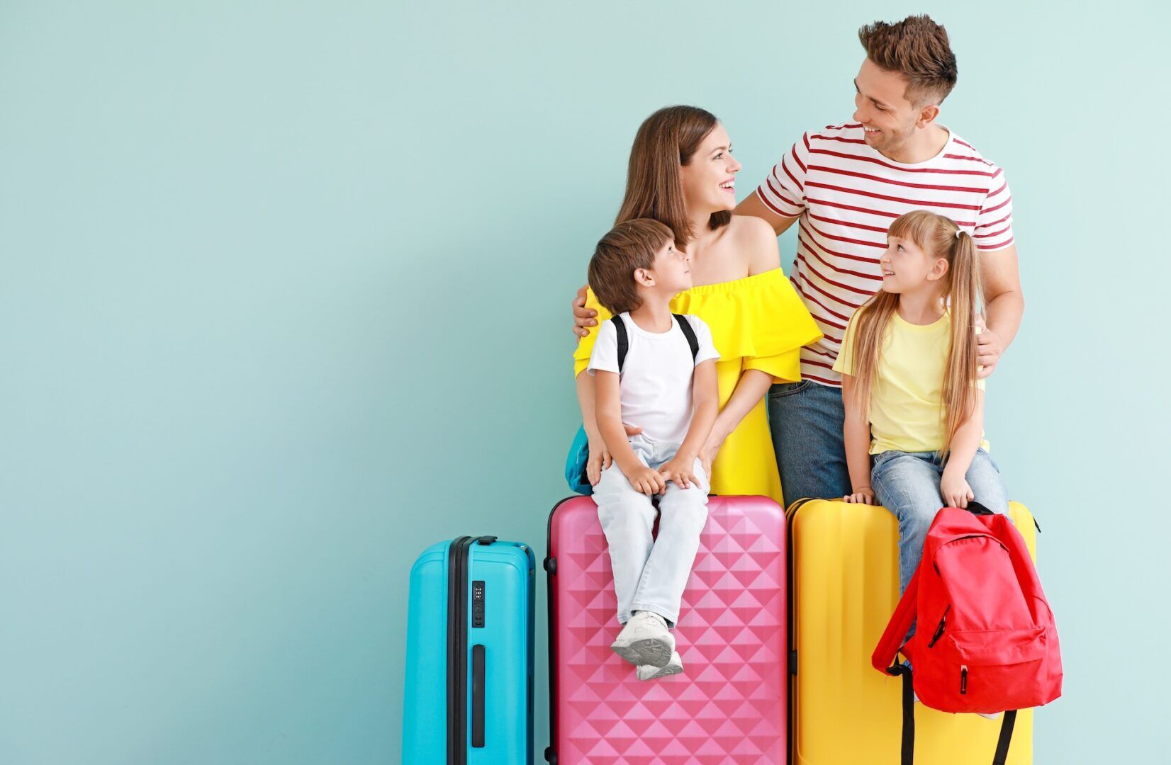 A smiling family of four sits on colorful suitcases against a light blue background. The parents and two children look at each other, appearing happy and ready for travel. The daughter holds a red backpack.