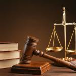 A wooden gavel resting on a block, three thick law books stacked, and a golden balance scale on a wooden table against a brown background.