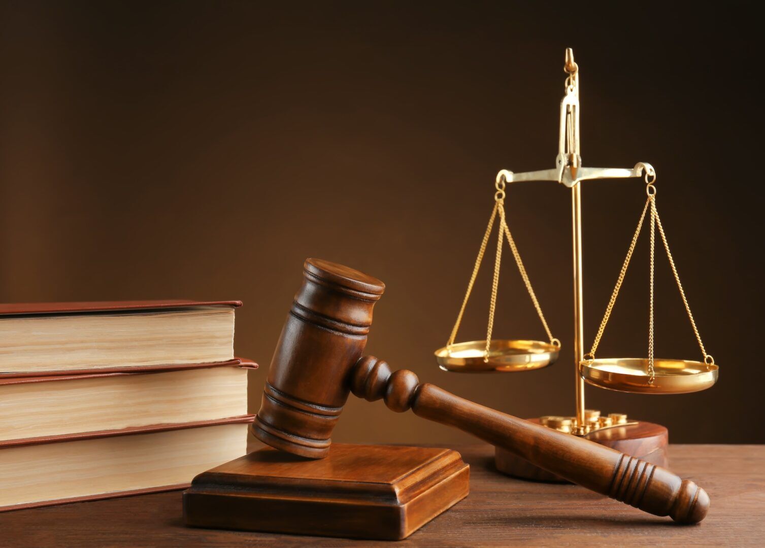 A wooden gavel resting on a block, three thick law books stacked, and a golden balance scale on a wooden table against a brown background.