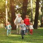 Two adults and three young children walk and run together through a sunny, green park with tall trees. The children are in front, smiling, while the adults follow behind, also smiling and enjoying the outdoors.