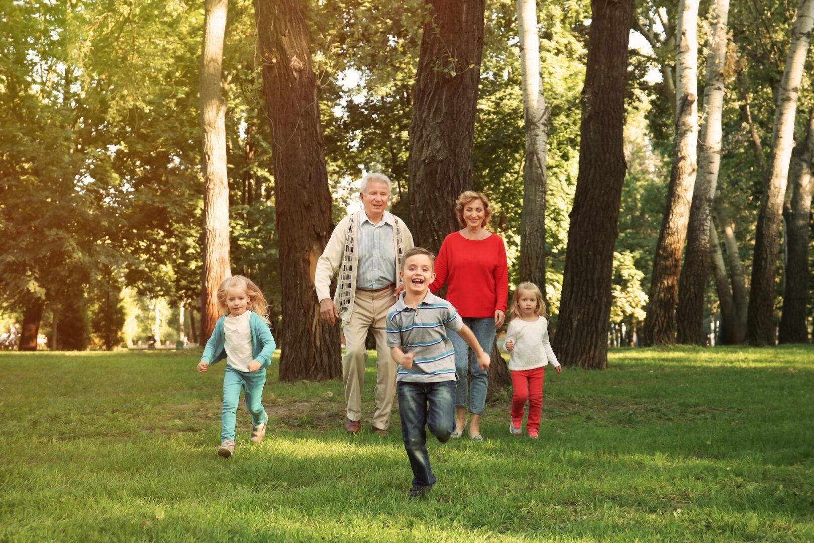 Two adults and three young children walk and run together through a sunny, green park with tall trees. The children are in front, smiling, while the adults follow behind, also smiling and enjoying the outdoors.