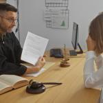 A judge in a robe shows legal documents to a woman sitting across a desk, who holds her head in her hand, appearing stressed. Legal books, a gavel, and scales of justice are on the desk.