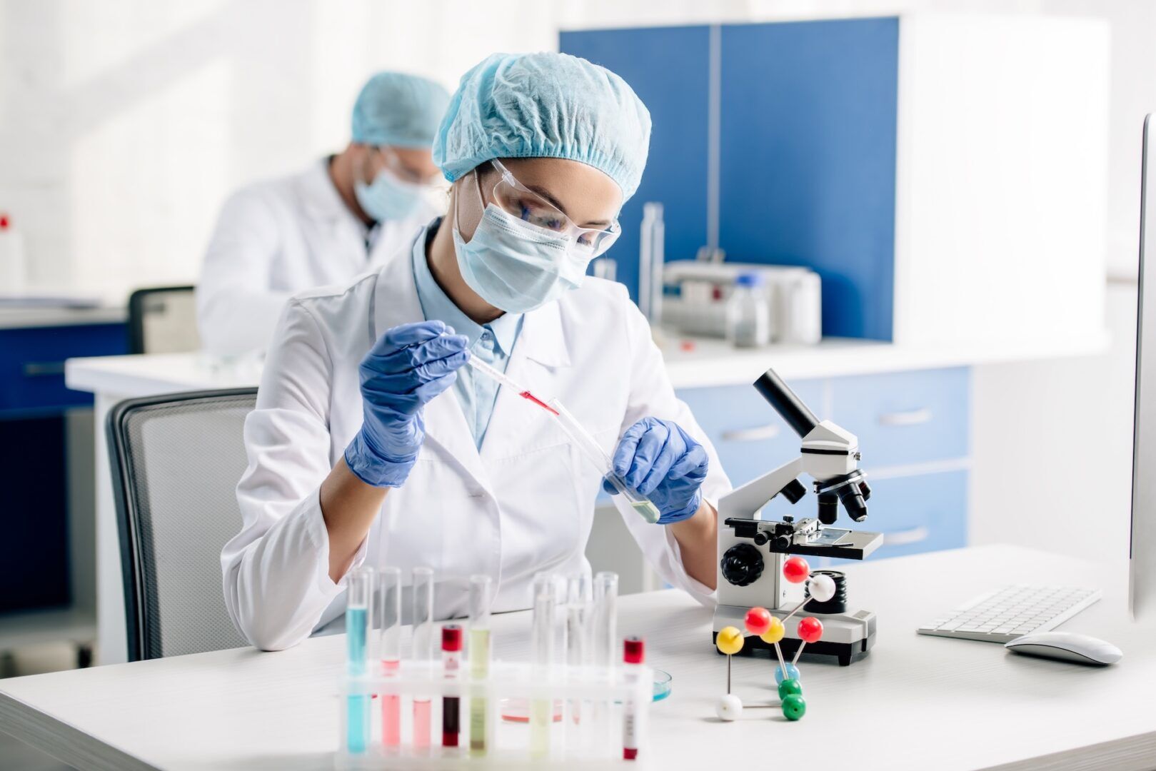 A scientist in protective gear uses a pipette to transfer liquid between test tubes in a laboratory, with a microscope and colorful samples on the desk. Another scientist works in the background.