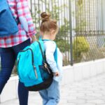 An adult and a young child with backpacks walk hand-in-hand on a sidewalk next to a fence. The child wears a blue backpack and light clothing, and the adult is dressed in jeans and a plaid shirt.