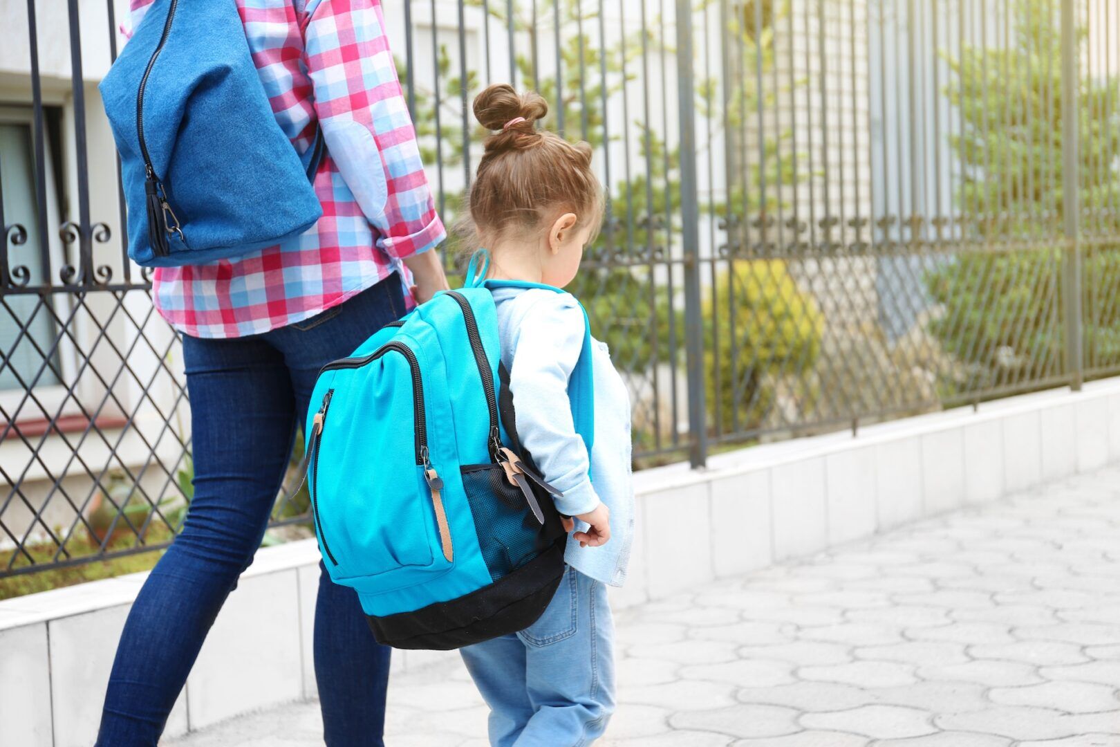 An adult and a young child with backpacks walk hand-in-hand on a sidewalk next to a fence. The child wears a blue backpack and light clothing, and the adult is dressed in jeans and a plaid shirt.