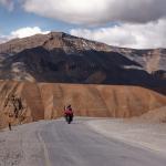 A person rides a motorcycle on a winding mountain road under a partly cloudy sky, with rugged, brown and rocky mountains in the background.