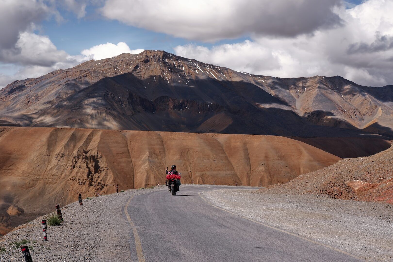 A person rides a motorcycle on a winding mountain road under a partly cloudy sky, with rugged, brown and rocky mountains in the background.