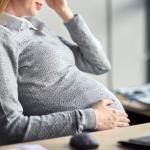 A pregnant woman sits at a desk, wearing a gray sweater and white collared shirt, resting one hand on her belly and the other on her forehead, appearing thoughtful or tired.