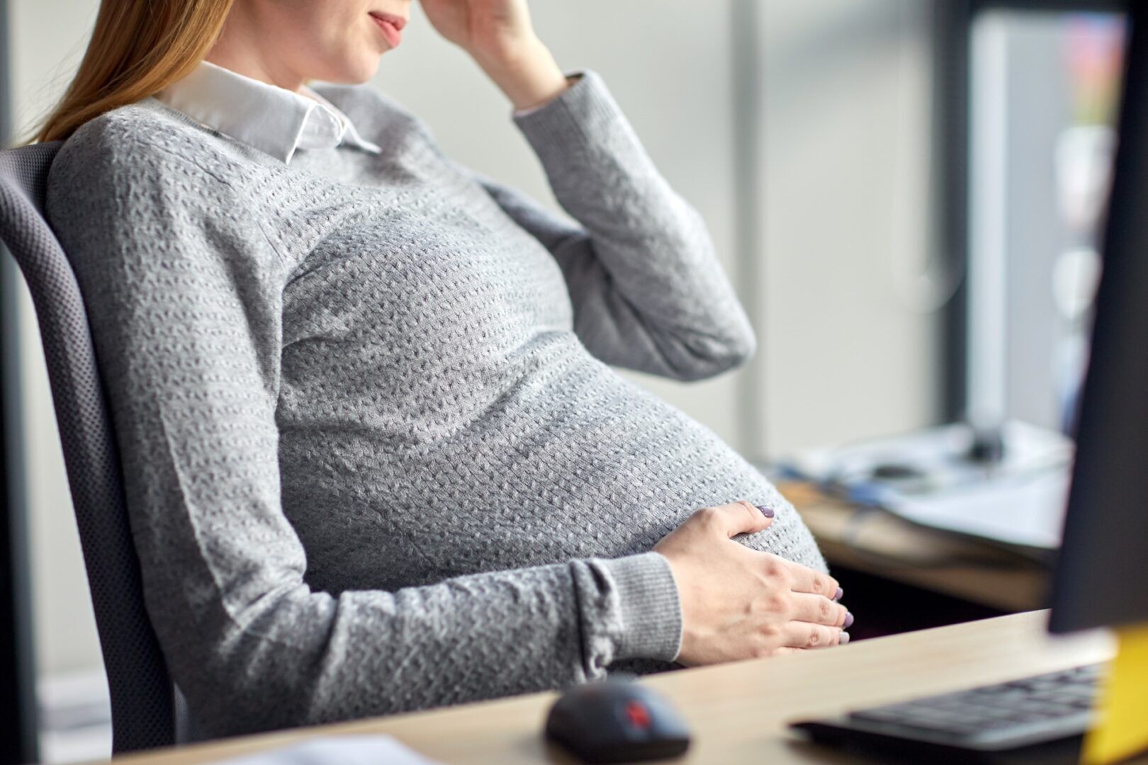 A pregnant woman sits at a desk, wearing a gray sweater and white collared shirt, resting one hand on her belly and the other on her forehead, appearing thoughtful or tired.