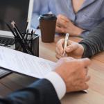 A person in business attire holds a clipboard with documents, while another person prepares to sign it with a pen. A laptop, pencil holder, and coffee cup are on the wooden desk.