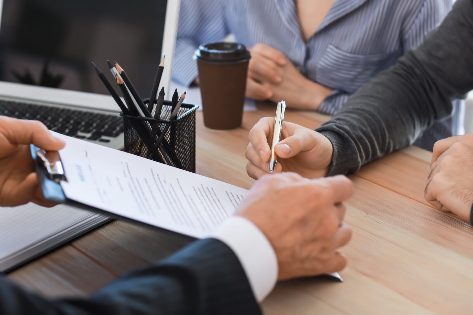 A person in business attire holds a clipboard with documents, while another person prepares to sign it with a pen. A laptop, pencil holder, and coffee cup are on the wooden desk.