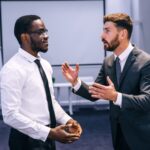 Two men in business attire have a serious discussion in an office setting. One man gestures with his hands while speaking, while the other listens attentively. A projector screen and flipchart are visible in the background.