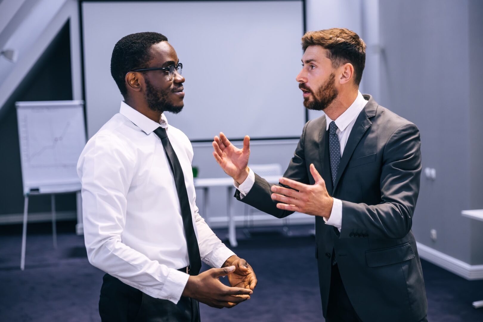 Two men in business attire have a serious discussion in an office setting. One man gestures with his hands while speaking, while the other listens attentively. A projector screen and flipchart are visible in the background.
