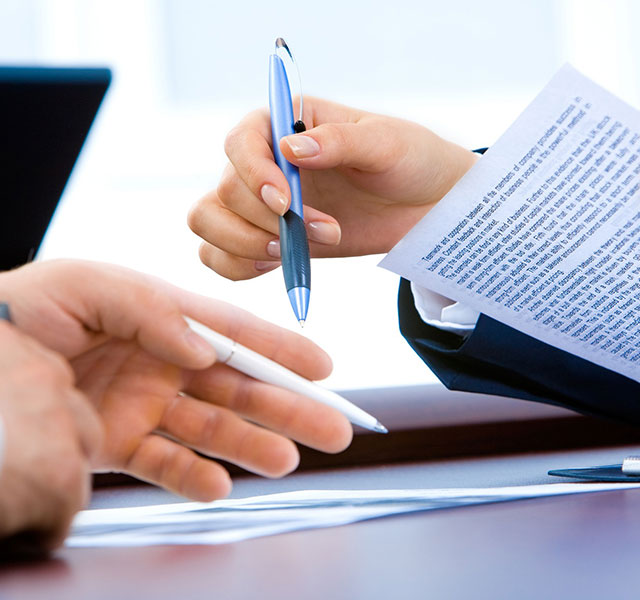 Two people sit at a desk, one holding a printed document and a pen, while the other extends a hand with a pen. A laptop and papers are visible on the table, suggesting a business meeting or document review.