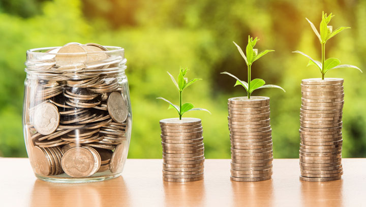 A glass jar full of coins sits beside three stacks of coins, each topped with a small green plant, symbolizing financial growth and investment. The background is blurred greenery.
