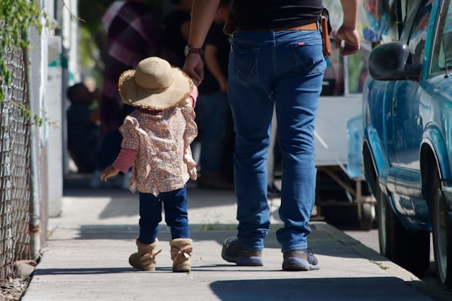 A small child wearing a sun hat and boots holds hands with an adult as they walk down a sunny sidewalk next to a blue car. The image is taken from behind.