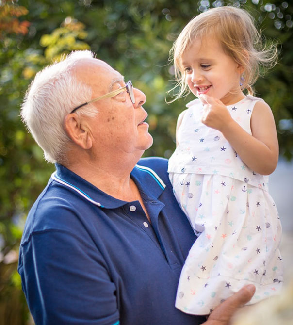 An elderly man in glasses and a blue shirt holds a smiling young girl in a white dress with star patterns. They look at each other happily outdoors with greenery in the background.