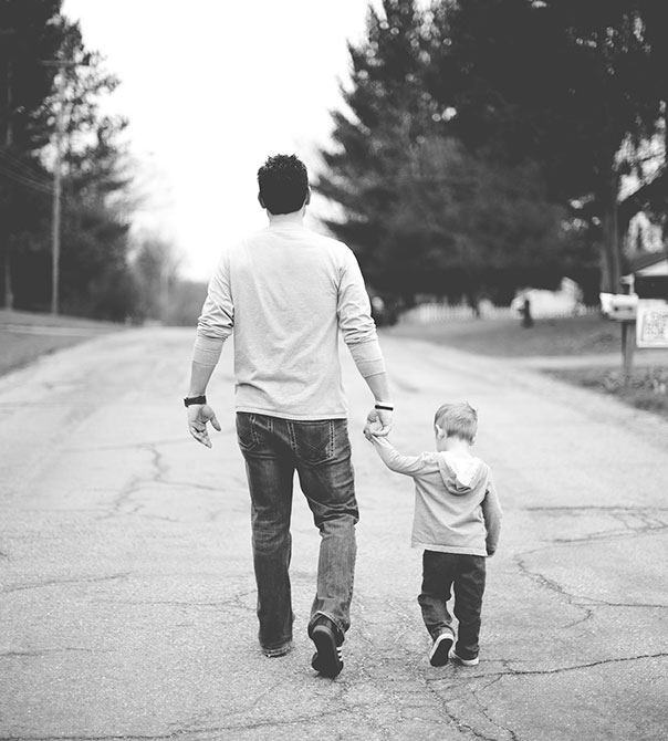 A man and a young boy walk hand in hand down the middle of a quiet street, photographed from behind in black and white. Trees and houses line the road on a cloudy day.