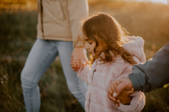 A young girl in a pink furry jacket holds hands with two adults while walking outdoors on a grassy path in warm, golden sunlight. The scene captures a sense of family and togetherness.