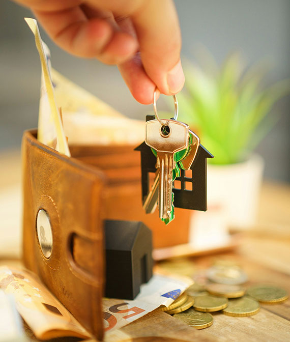 A hand holds house-shaped keychains with keys near an open wallet filled with euro banknotes, coins, and a small house model, with a green plant in the background.