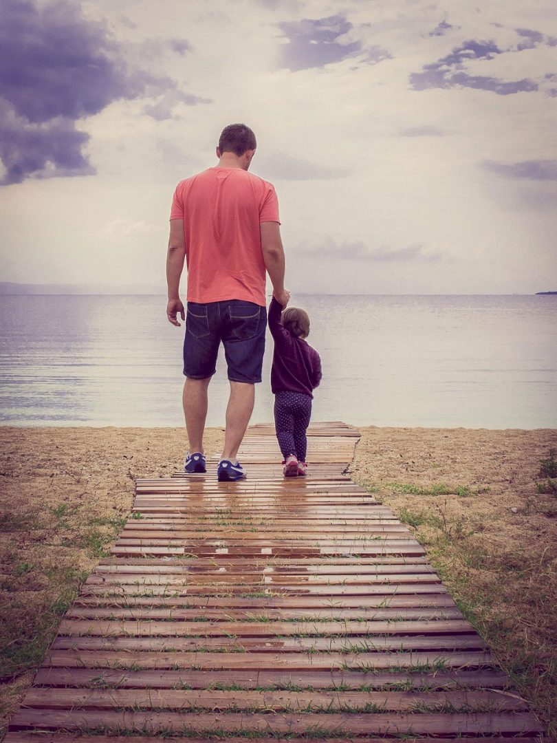 A man and a small child walk hand in hand on a wooden path leading to a sandy beach, with calm water and a cloudy sky in the background. Both are seen from behind.