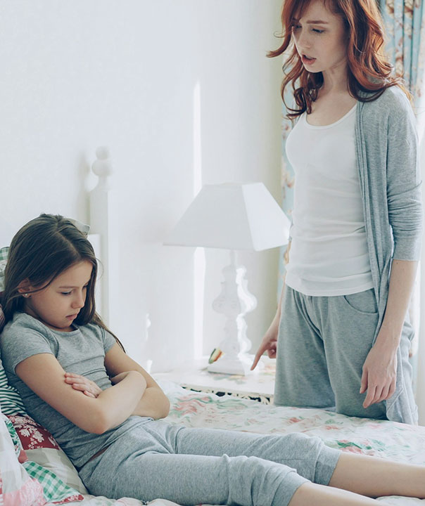 A woman stands near a bed, looking upset and pointing while a young girl sits on the bed with her arms crossed, appearing sad and looking down. Both are wearing gray clothes in a softly lit bedroom.