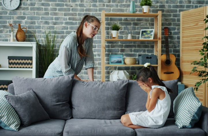 A woman leans over a gray couch, speaking to a young girl who is sitting and covering her face with her hands, appearing upset. The room has modern decor, shelves, plants, and a guitar in the background.