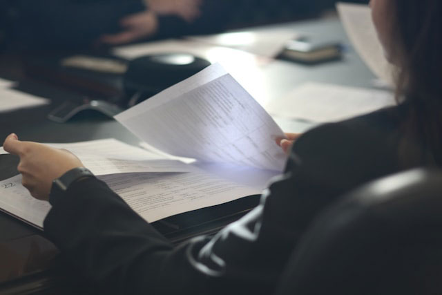 Person in professional attire holding and reviewing a stack of documents at a table, with other papers, a gavel, and another person in the background, suggesting an office or meeting setting.