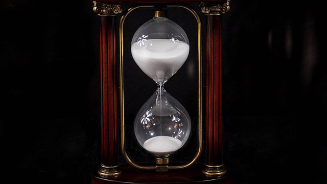 A glass hourglass with white sand is set within a dark wooden frame with brass accents, against a black background.