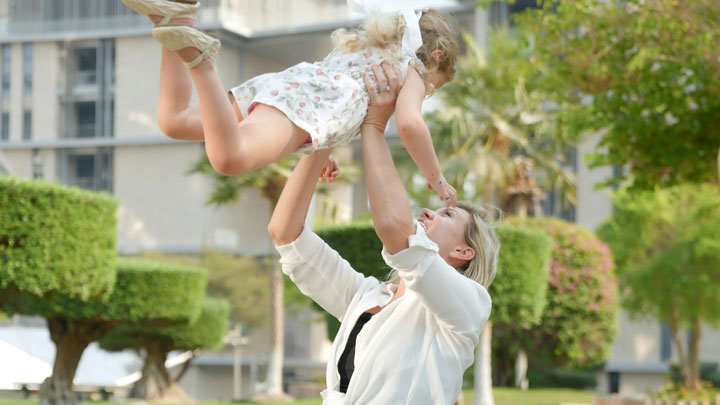 A woman joyfully lifts a young girl in a floral dress above her head outdoors, both smiling with trees, shrubs, and buildings in the background.