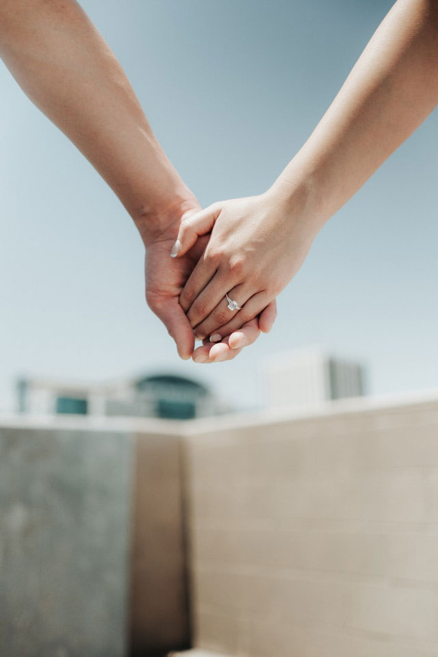 Two people holding hands outdoors, with one wearing an engagement ring. The background is blurred, showing a blue sky and distant buildings.
