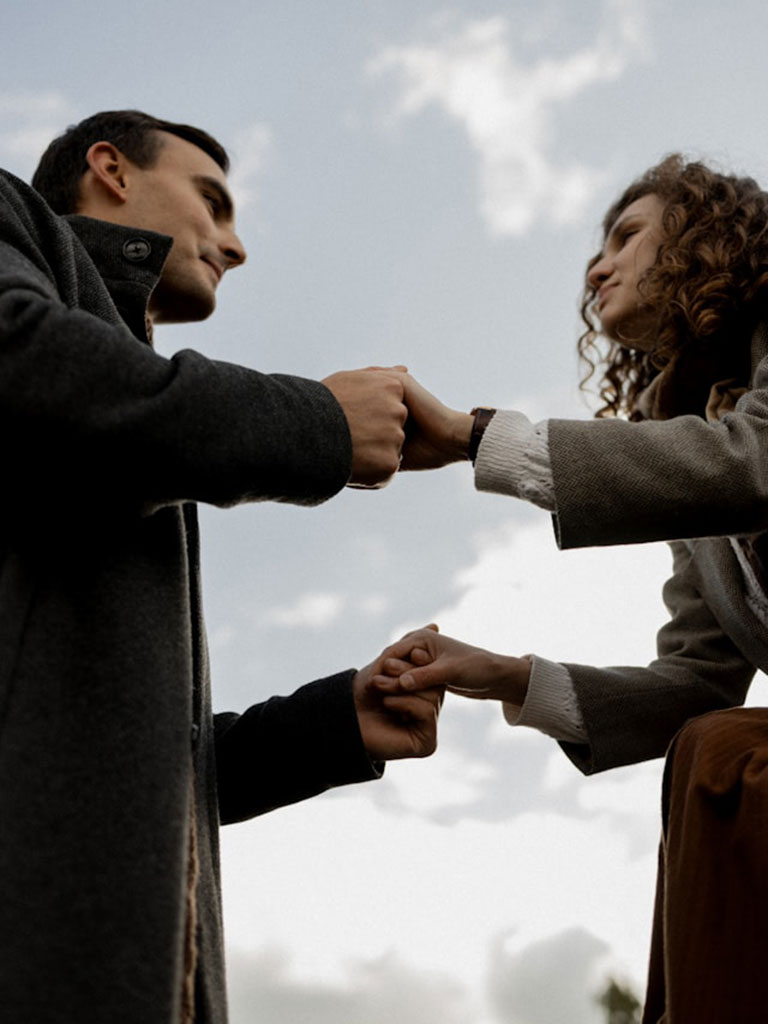 Two people wearing coats stand outdoors against a cloudy sky, facing each other and gently holding hands, forming a close and intimate connection. The photo is taken from a low angle.