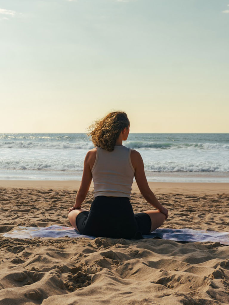 A person with curly hair sits cross-legged on a sandy beach, facing the ocean and horizon, with calm waves and a clear sky in the background.