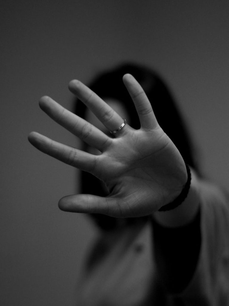 A person with long hair holds an open hand toward the camera, obscuring their face. The image is in black and white, and the person's ring is visible on their finger.