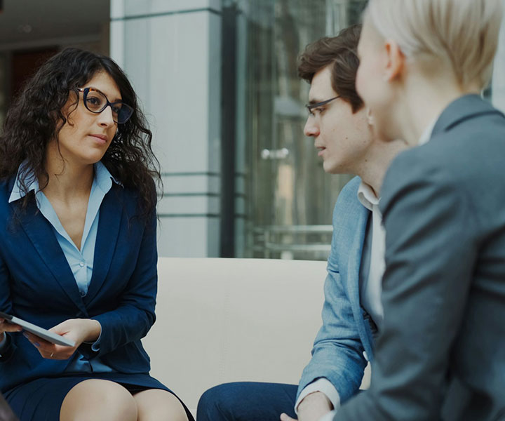 Three people in business attire sit together indoors, engaged in a serious conversation. One woman with curly hair and glasses holds a tablet, while the other two, a man and a woman, listen attentively.