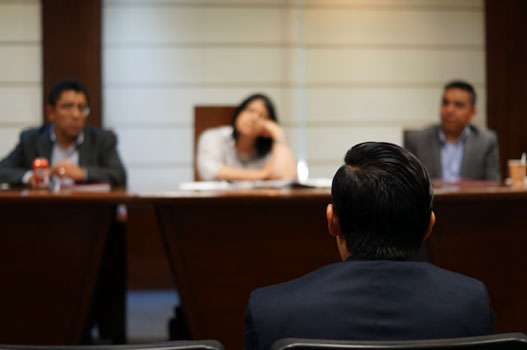 A person in a suit sits facing three people across a table in a formal meeting or interview setting. The three people appear to be listening attentively to the person in the foreground.