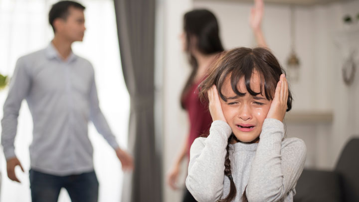 A young girl covers her ears and cries while two adults argue in the background of a home setting. The scene suggests family conflict and distress.