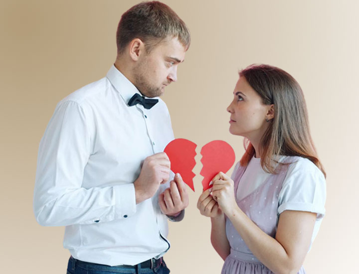 A man and a woman face each other holding two halves of a broken red paper heart. They both have serious expressions and are standing against a plain beige background.