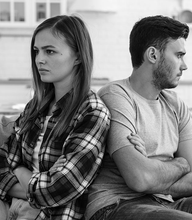 A young woman and man sit back-to-back on a couch with arms crossed, both looking upset and facing away from each other. The mood appears tense and they seem to be having a disagreement.