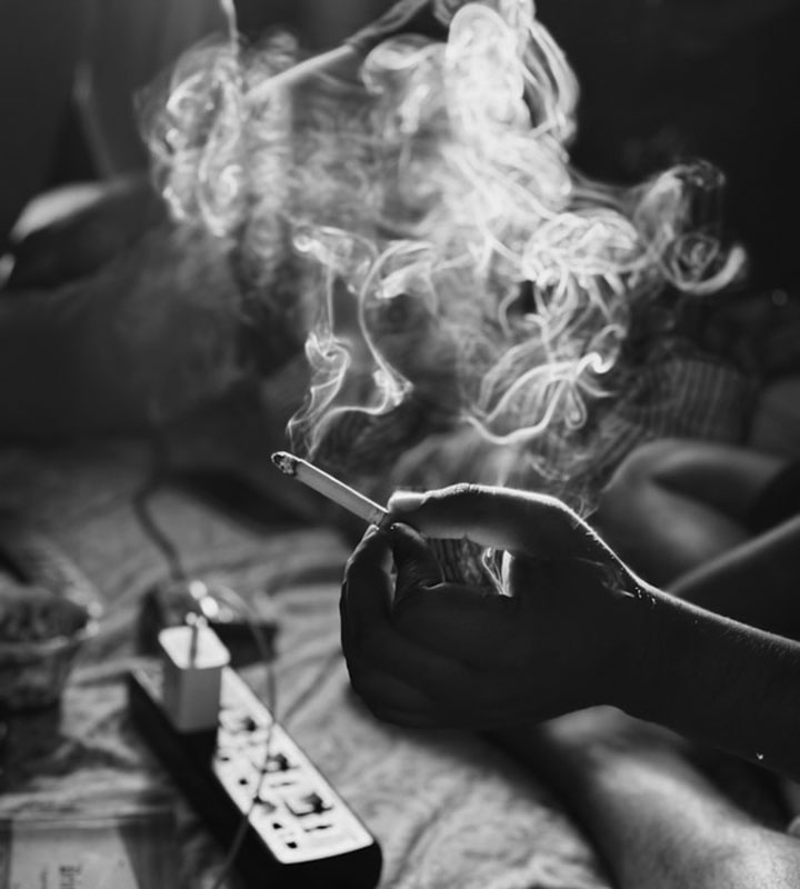 A hand holding a lit cigarette with swirling smoke, captured in black and white. An extension cord and other objects are visible on a table, and people sit in the blurred background.