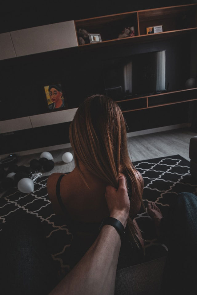 A person with long brown hair sits on a black and white patterned rug, facing away from the camera, while another person's hand gently holds their hair. Black and white balloons are scattered on the floor nearby.