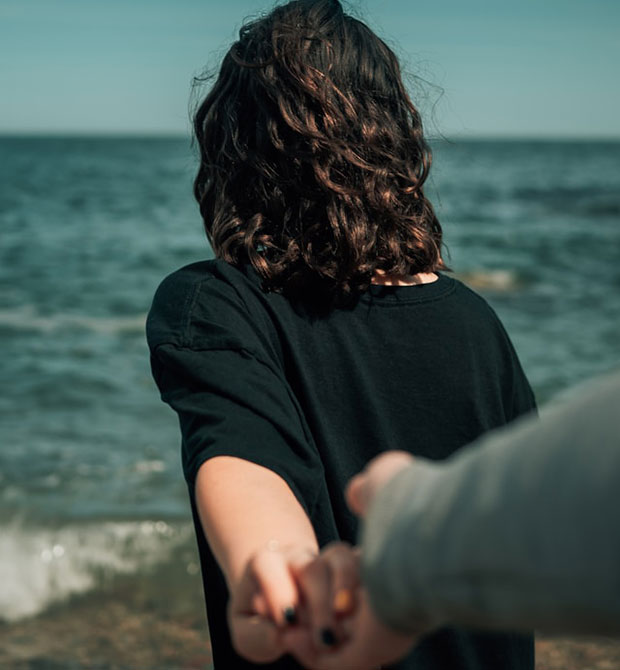A person with wavy brown hair, wearing a black shirt, stands facing the ocean while holding another person’s hand, leading them forward. The scene is set on a sunny day by the water.