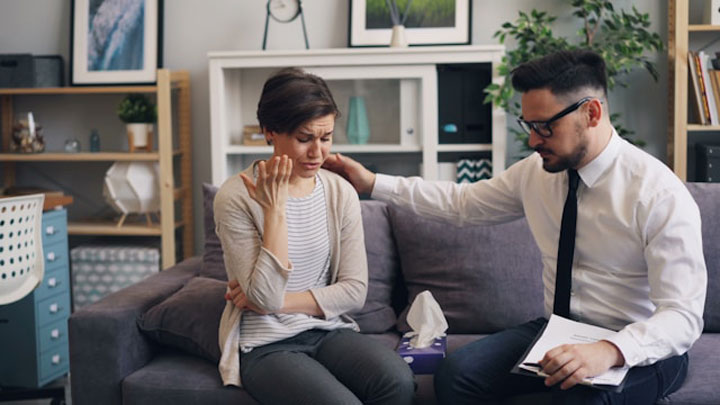 A woman sits on a couch looking upset while a man beside her, holding a notebook, gently rests his hand on her shoulder in a comforting gesture. They appear to be in a counseling or therapy session.
