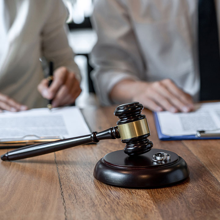 A judge's gavel and wedding rings rest on a wooden table, with two people in the background reviewing and signing legal documents.